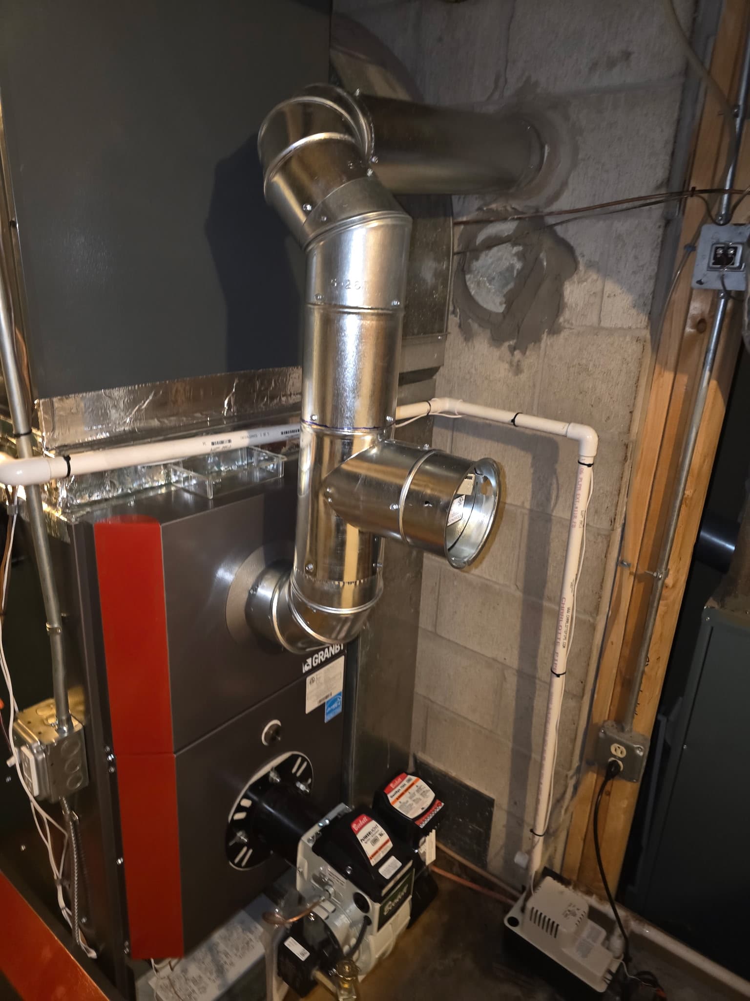 Residential furnace with silver metal exhaust pipes and white PVC piping against a concrete wall.
