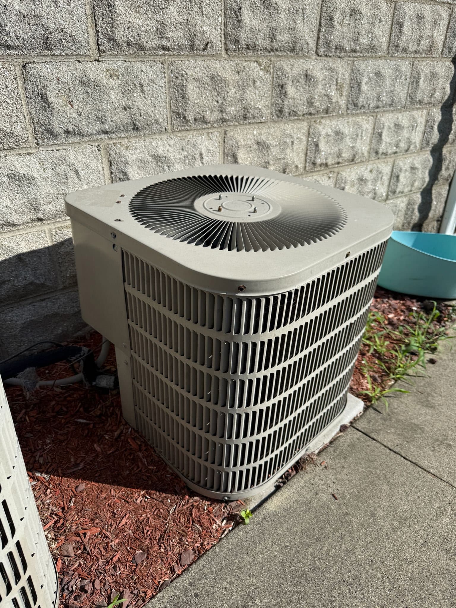 Outdoor central air conditioning condenser unit positioned against a light grey stone block wall.