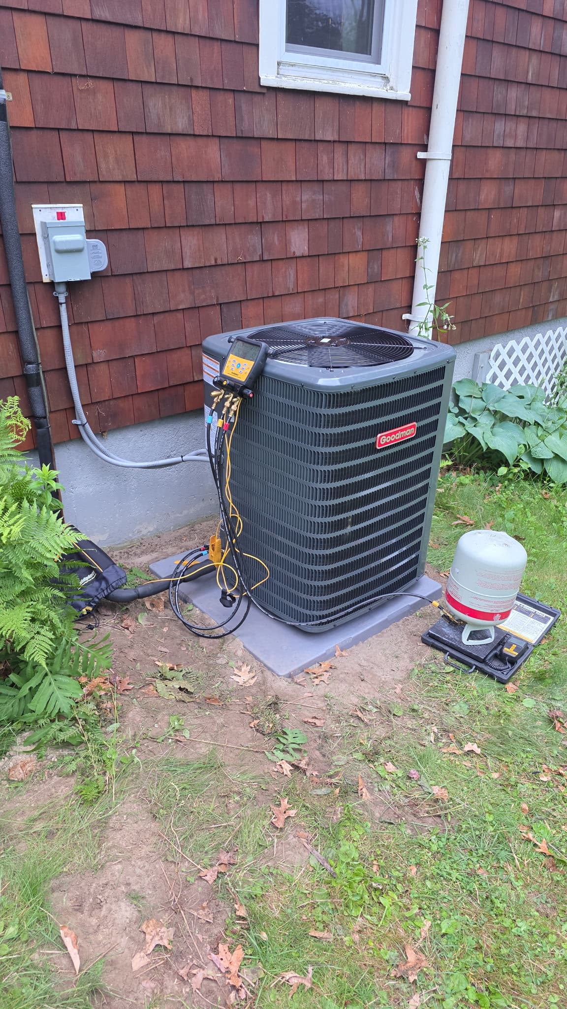 Goodman air conditioning unit being serviced with diagnostic gauges and refrigerant tank on scale.