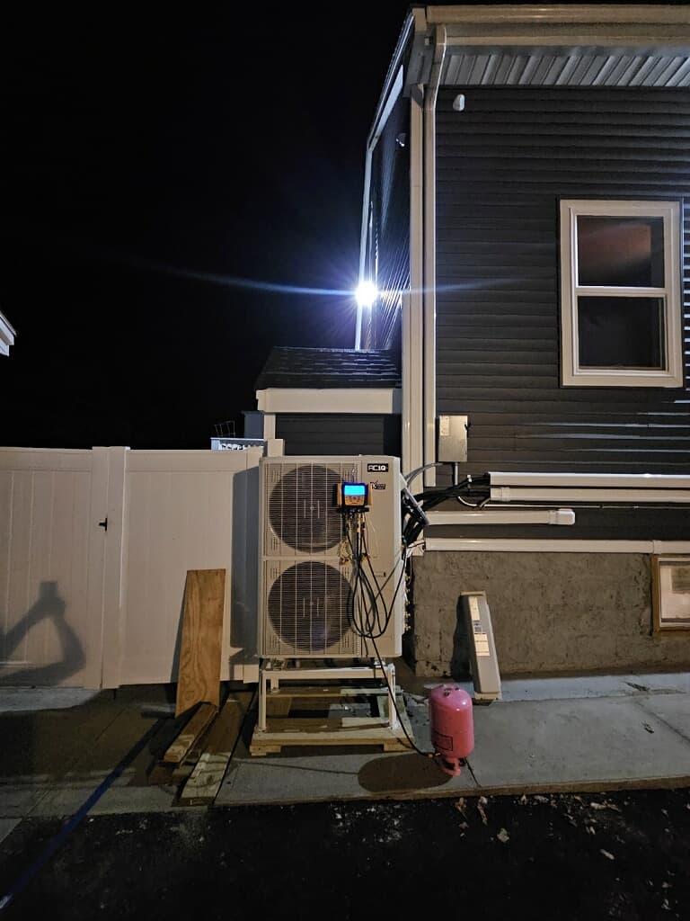 Outdoor HVAC unit being serviced at night with a digital manifold and pink refrigerant tank.