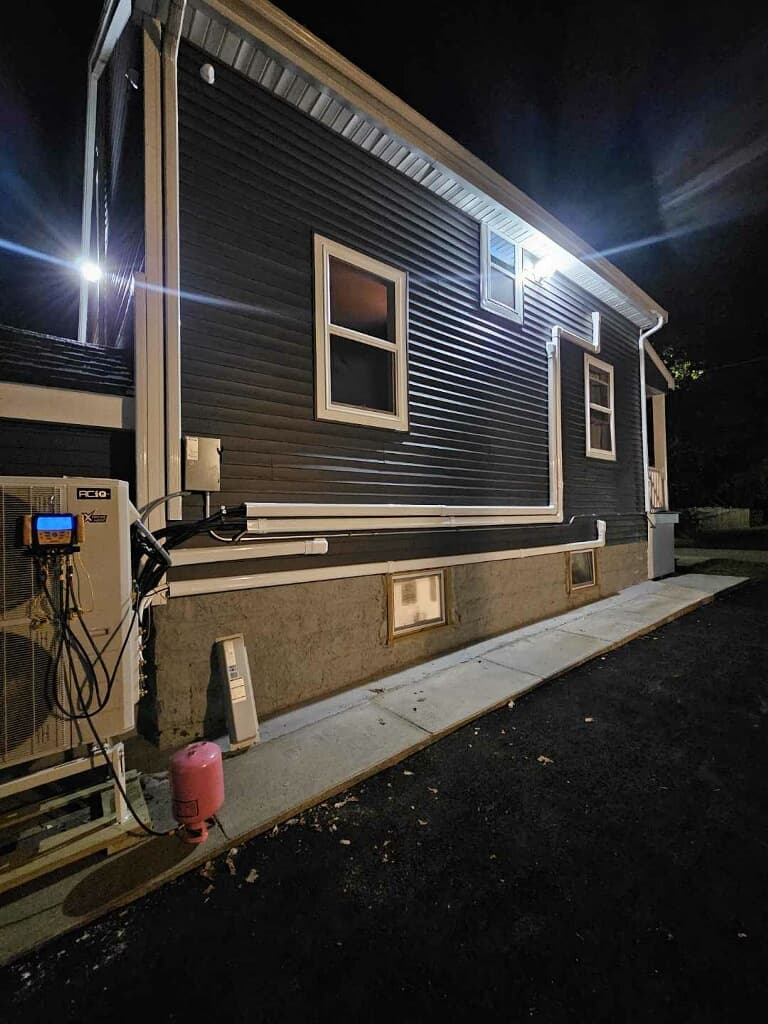 Heat pump unit installed on dark siding at night with white conduits and pink tank.